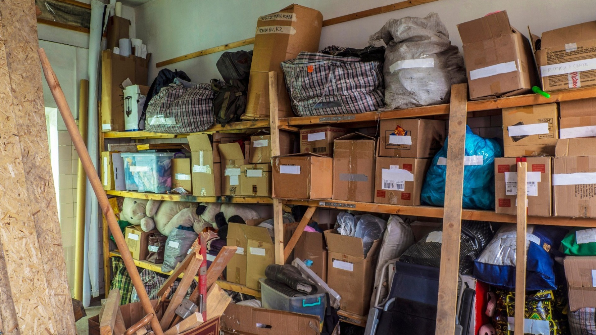 A cluttered, messy wooden shelf in a storage area is filled with disorganized cardboard boxes, plastic bags, plaid storage bags, and various loose items, reflecting a busy or untidy environment.
