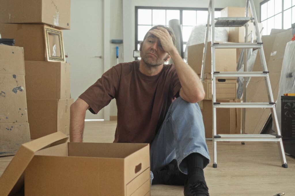 Man sitting on floor with hand to forehead beside an open cardboard box in a building; fatigue packing transition.